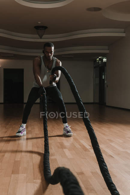 Bonito afro-americano masculino realizando exercício com cordas enquanto exercitando-se em quarto espaçoso do ginásio moderno — Fotografia de Stock