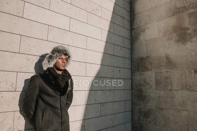 Afrikanisch-amerikanischer Mann mit Pelzmütze lehnt an Ziegelmauer an sonnigem Tag auf der Stadtstraße — Stockfoto