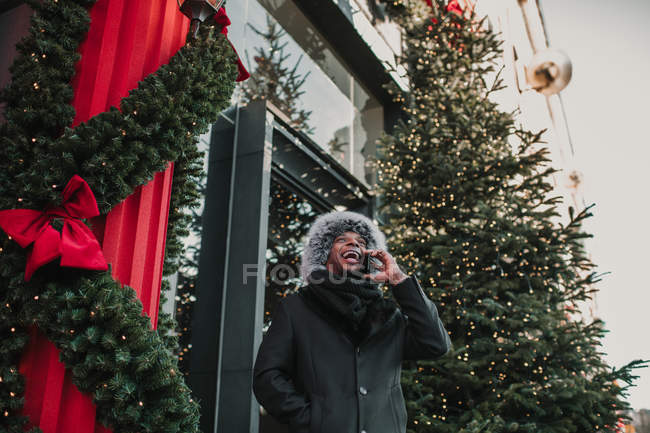Afrikanisch-amerikanischer Mann in warmer Kleidung spricht auf Smartphone, während er auf der Stadtstraße in der Nähe eines Gebäudes steht und einen weihnachtlich geschmückten Nadelbaum hat — Stockfoto