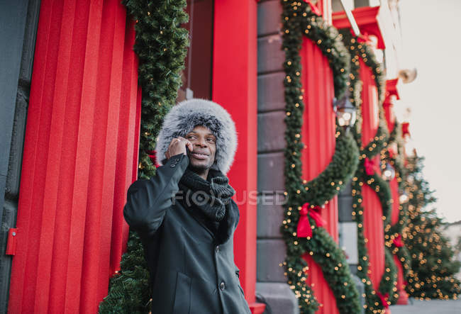 Афроамериканець у теплому одязі розмовляє на смартфоні, стоячи на вулиці біля будинку і хвойного дерева, прикрашеного Різдвом. — стокове фото