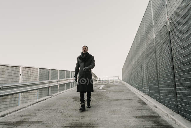 Hombre afroamericano en ropa de moda caminando sobre asfalto mojado cerca de valla en día gris en la ciudad - foto de stock