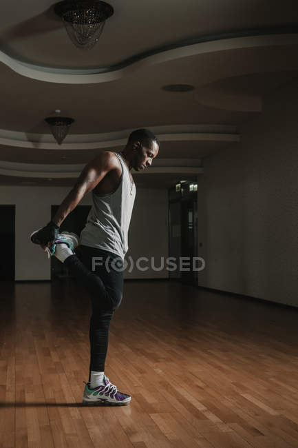 Vista lateral do cara negro atraente fazendo exercício de aquecimento para as pernas enquanto está em pé no quarto espaçoso do ginásio moderno — Fotografia de Stock