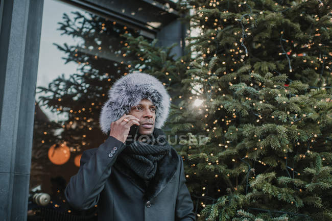 Hombre afroamericano en ropa de abrigo hablando en un teléfono inteligente mientras está de pie en la calle de la ciudad cerca del edificio y el árbol de coníferas decorado para Navidad - foto de stock