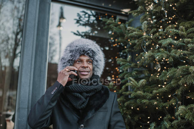 Excited African American man in warm clothes talking on smartphone while standing on city street near building and conifer tree decorated for Christmas — Stock Photo