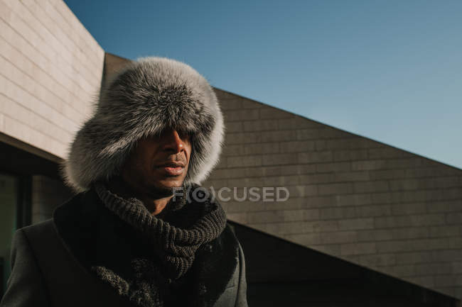 Привлекательный афроамериканец в меховой шапке стоит возле современного здания в солнечный день на городской улице — стоковое фото