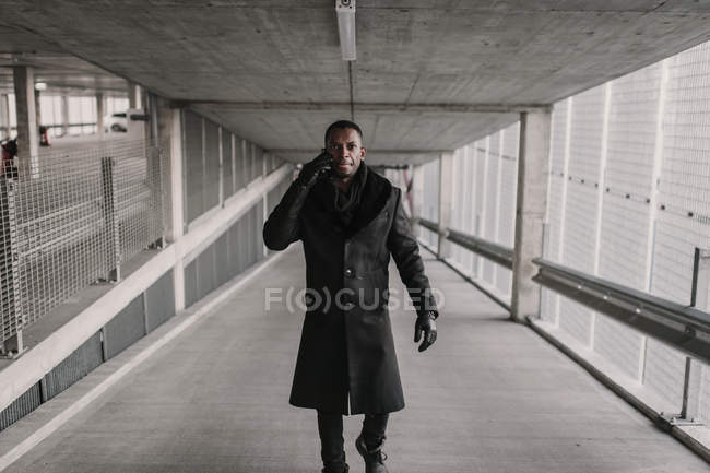 African American man in stylish outfit walking in passage of modern building and having smartphone conversation — Stock Photo