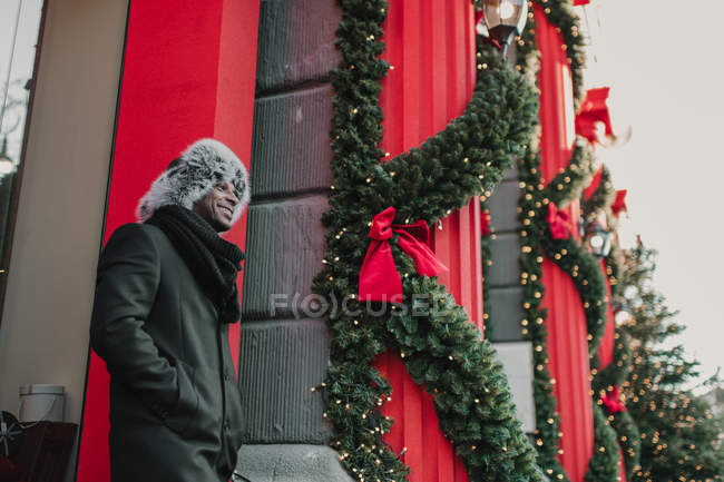 Афроамериканец в теплой одежде, стоя на городской улице рядом со зданием и хвойной елкой, украшенной на Рождество — стоковое фото