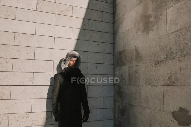 Attractive African American man in fur hat leaning on brick wall on sunny day on city street — Stock Photo