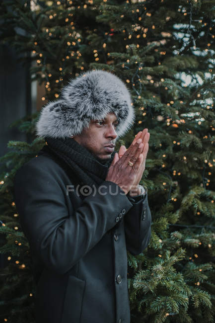 Handsome African American man in warm clothes rubbing hands while standing near Christmas tree with fairy lights on city street — Stock Photo