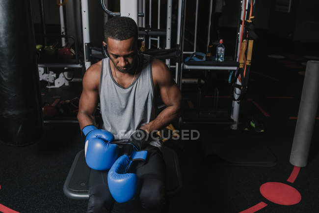 Hombre negro guapo sentado en la máquina de ejercicios y ponerse guantes de boxeo azul durante el entrenamiento en el gimnasio moderno - foto de stock