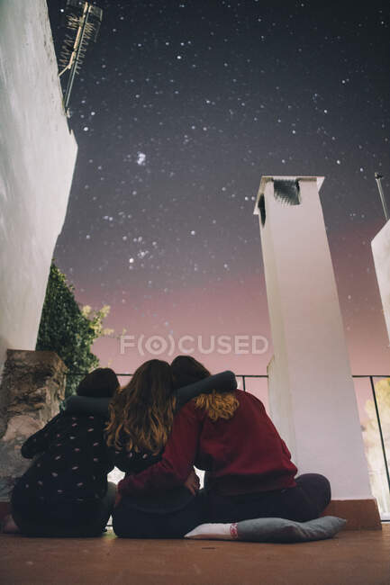 Rückansicht junger Freunde, die sich umarmen und den majestätischen Sternenhimmel bewundern, während sie nachts gemeinsam auf der Terrasse sitzen — Stockfoto