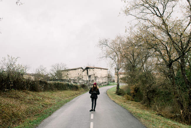 Юная леди в зимней одежде и шляпе с камерой и руками в карманах на сельской дороге в orduna, Испания — стоковое фото