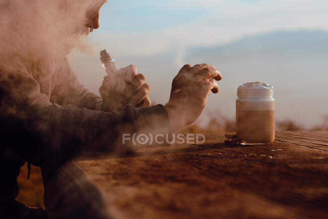 Unrecognizable person vaping and using smartphone to take photo of cup of fresh coffee while sitting at wooden table in countryside — Stock Photo
