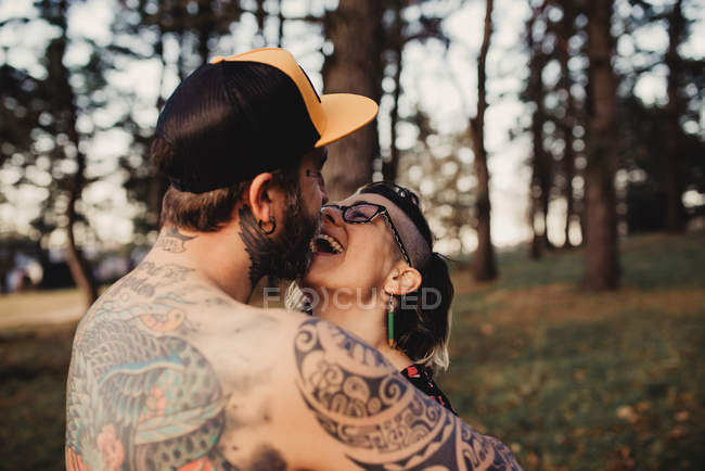 Vista posteriore del giovane uomo senza maglietta in tatuaggi con schiocco che abbraccia la donna nel parco su sfondo sfocato — Foto stock