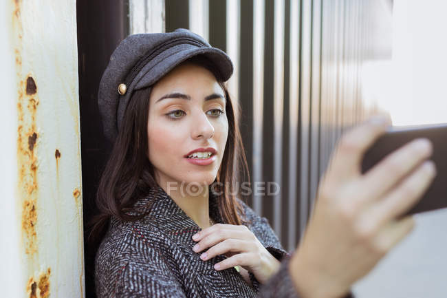 Encantadora dama hispana con abrigo y gorra tomando selfie cerca de la pared de metal - foto de stock