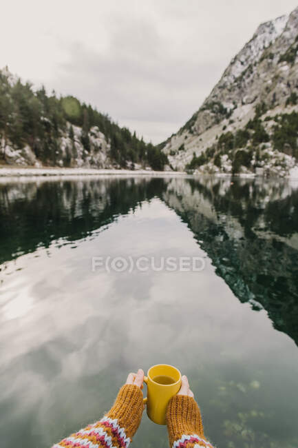 Mano de la cosecha humana sosteniendo taza amarilla cerca de la increíble vista de la superficie del agua entre las altas montañas con árboles en la nieve y el cielo nublado en los Pirineos - foto de stock