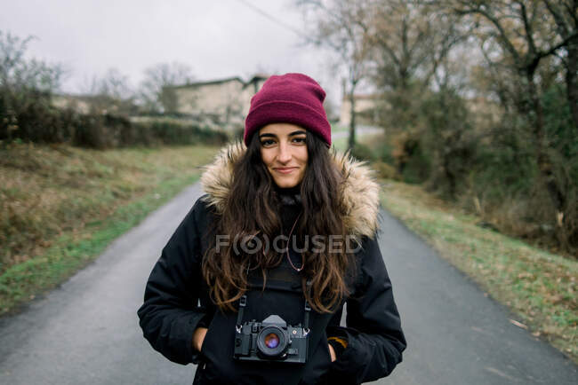 Attrayant jeune femme gaie en hiver porter et chapeau avec caméra et les mains dans les poches sur la route de campagne à Oruna, Espagne — Photo de stock