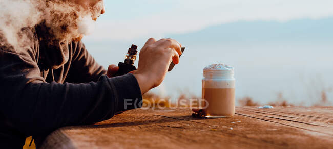 Persona irreconocible vapeando y utilizando el teléfono inteligente para tomar una foto de la taza de café fresco mientras está sentado en la mesa de madera en el campo - foto de stock