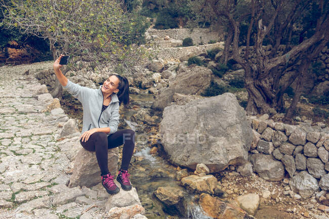 Bella donna in abito casual allegramente sorridente e utilizzando smartphone moderno per scattare selfie mentre seduto sulla roccia vicino al torrente con acqua dolce pulita in un parco maestoso — Foto stock