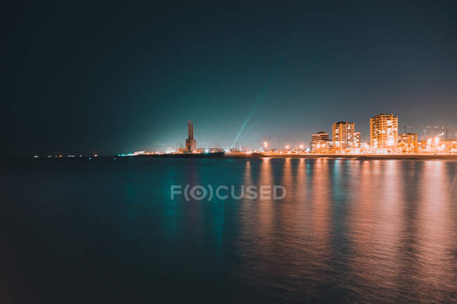 Superfície de água do mar tranquila perto da cidade costeira brilhantemente iluminada na bela noite — Fotografia de Stock