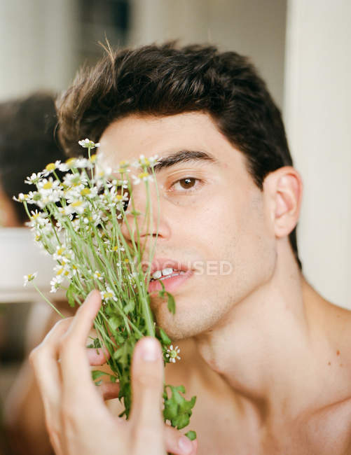 Боковой вид молодого парня без рубашки со свежими белыми цветами в руках, смотрящего в камеру на размытом фоне — стоковое фото