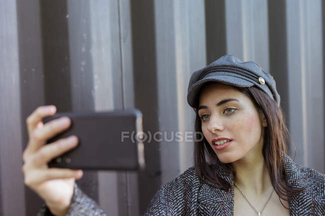 Charmosa senhora hispânica de casaco e boné tirando selfie perto da parede de metal — Fotografia de Stock