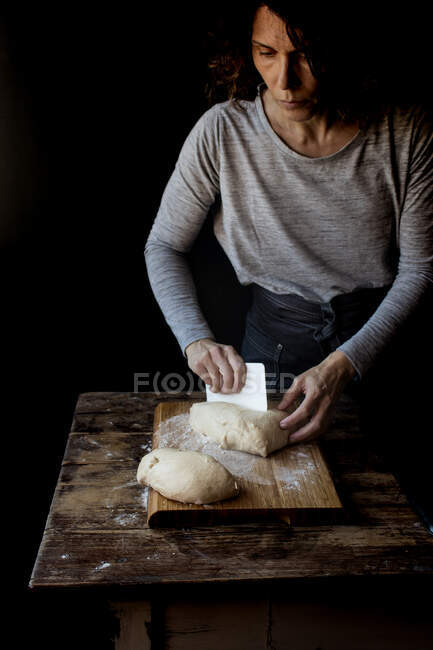 Cultivo humano celebración de la masa con harina sobre tabla de cortar cerca de mesa de madera sobre fondo negro - foto de stock