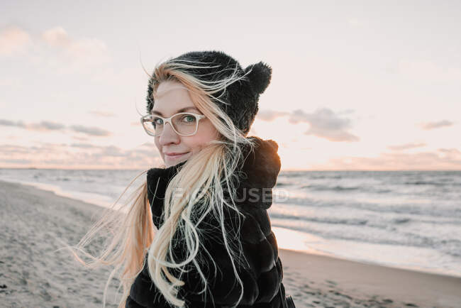 Вид сзади улыбающейся привлекательной дамы в очках и шубе, прогуливающейся по берегу у воды на закате в Литуании — стоковое фото