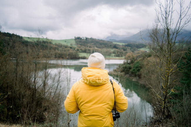 Back view of human in yellow coat and hat with camera looking at picturesque view of lake between hilss in Orduna, Spain - foto de stock