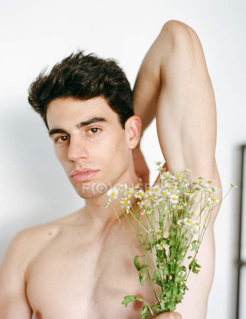 Side view of young shirtless guy with fresh white flowers in hands looking at camera on blurred background — Stock Photo