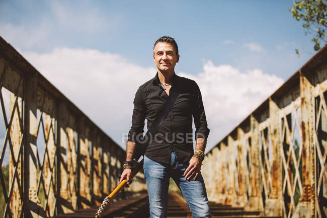 Hombre adulto con guitarra eléctrica de pie en el puente resistido y mirando a la cámara en el día soleado en el campo - foto de stock