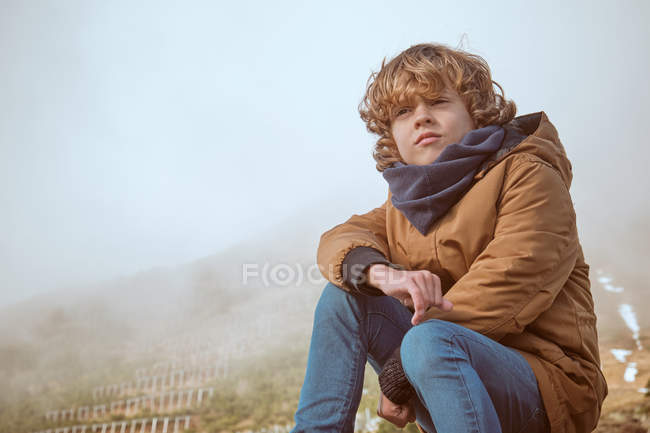 Adorable boy in warm clothes looking away while sitting on background of majestic hill on misty day in countryside — Stock Photo
