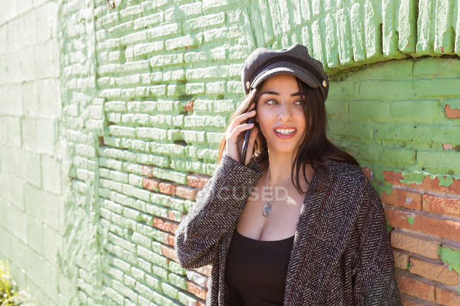 Smiling Hispanic young woman talking on mobile phone near green brick wall — Stock Photo