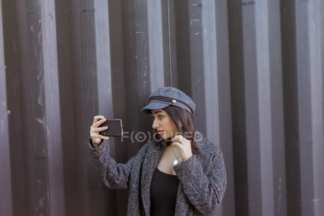 Stylish young woman in coat and cap posing and taking selfie near metal wall — Stock Photo