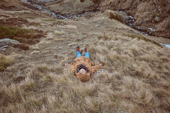 Enfant en vêtements chauds couché sur de l'herbe sèche sur la pente d'une colline près d'un petit ruisseau à la campagne automnale — Photo de stock