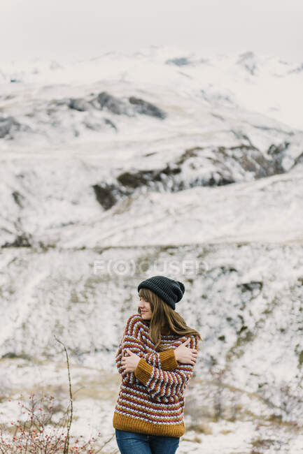 Menina atraente em suéter e chapéu com as mãos cruzadas perto da colina no fundo borrado em Pirinéus — Fotografia de Stock
