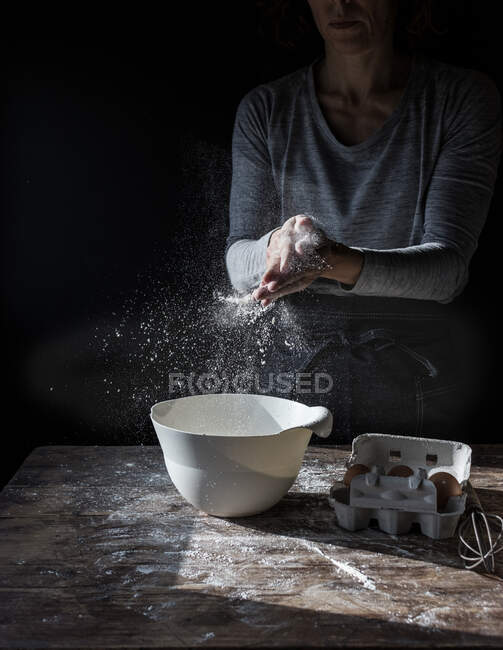 Crop lady clapping hands in flour near bowl, box of eggs and whisk on wooden table on black background — Stock Photo