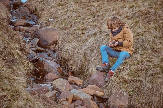 Sweet boy in warm clothes sitting on dry grass near creek with clean water in autumn countryside — Stock Photo