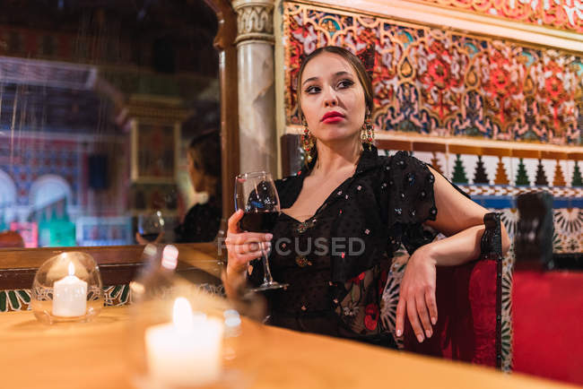 Attractive young woman in dress sitting at table near glass of drink and burning candles in luxury room of cafe decorated by mosaic — Stock Photo