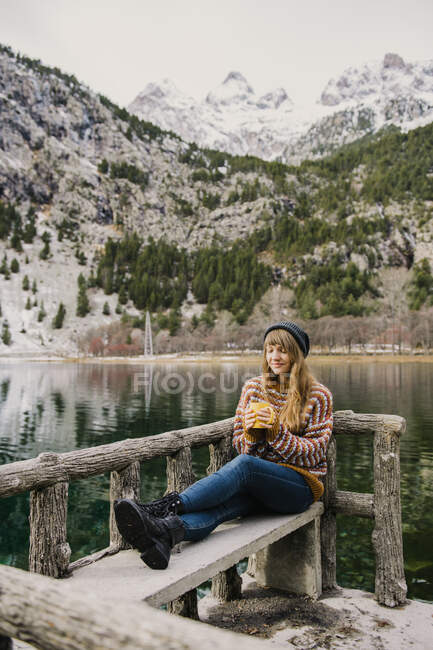 Молода пані сидить на лавці заплющених очей біля дивовижної поверхні води між високими горами з деревами у снігу в Піренеях. — стокове фото