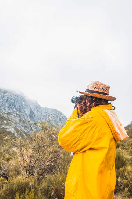 Vista lateral da senhora de chapéu e capa de chuva amarela disparando montanhas com nuvens na câmera em Isoba, Castela e Leão, Espanha — Fotografia de Stock