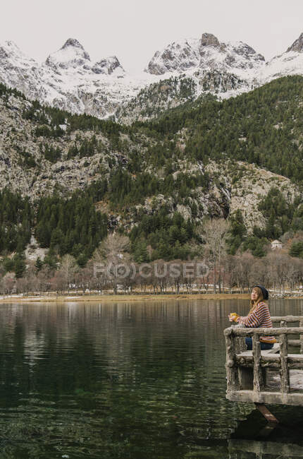 Vista lateral de la señora sentada en el banco y mirando la increíble vista de la superficie del agua entre altas montañas con árboles en la nieve y el cielo nublado en los Pirineos - foto de stock