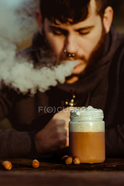 Homem vaping perto de uma xícara de café fresco enquanto sentado à mesa de madeira no campo — Fotografia de Stock