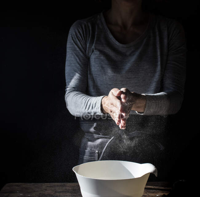 Señora de la cosecha aplaudiendo las manos en la harina cerca de cuenco, caja de huevos y batidor en la mesa de madera sobre fondo negro - foto de stock