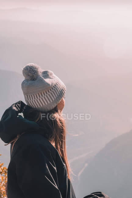 Side view of young lady in coat and hat looking on valley from top of mountain and cloudy sky — Stock Photo