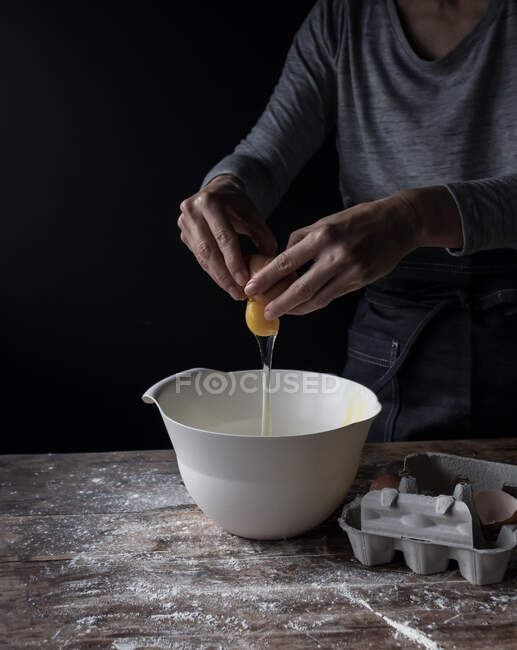 Crop human breaking egg in bowl on wooden table with flour on black background — Stock Photo
