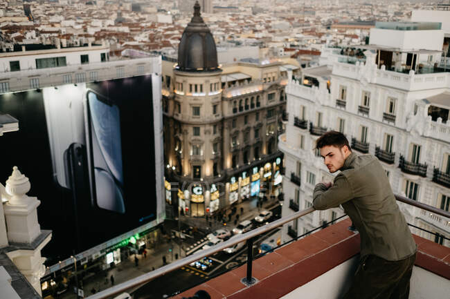 Visão traseira de cara elegante jovem em desgaste casual perto de trilhos no telhado olhando para o fundo da paisagem urbana em Madrid, Espanha — Fotografia de Stock