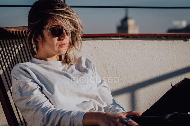 Giovane signora elegante in abbigliamento casual e occhiali da sole seduto sulla sedia sul tetto e cielo blu con la cima della torre a Madrid, Spagna — Foto stock
