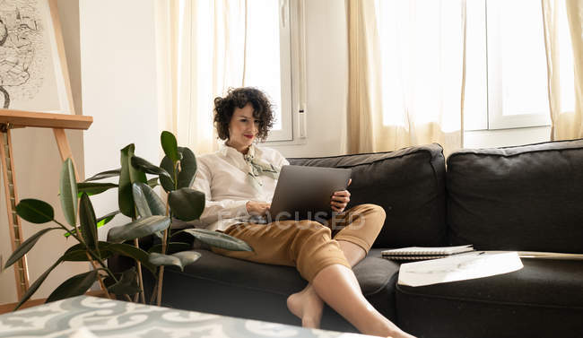 Junge attraktive, glückliche Frau sitzt auf einem Sofa und benutzt Laptop in der Nähe von Farben auf Bettwäsche im hellen Raum — Stockfoto