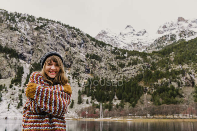 Attraktive junge Frau in Pullover und Hut mit verschränkten Händen in der Nähe eines Hügels vor verschwommenem Hintergrund in den Pyrenäen — Stockfoto